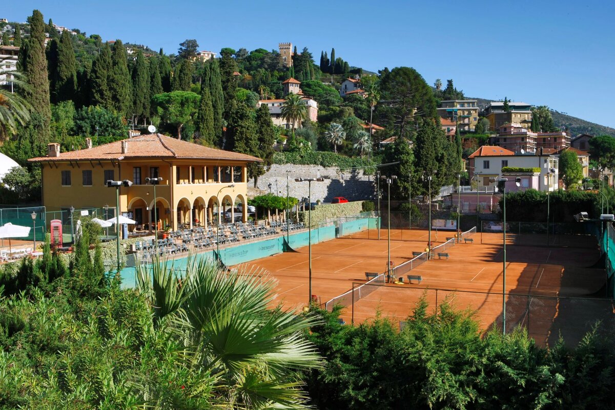 Clay tennis courts with building and greenery in the background