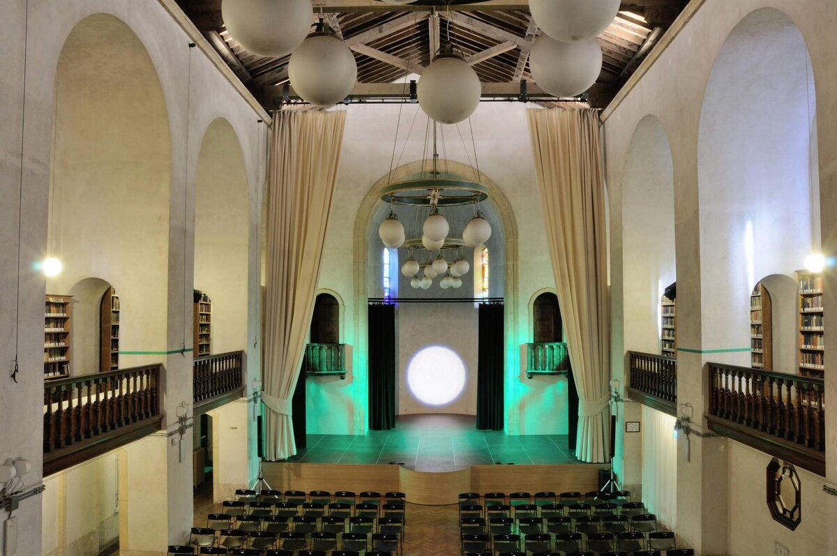 Interno di un teatro con palco, sedie e luci soffuse in un edificio storico.